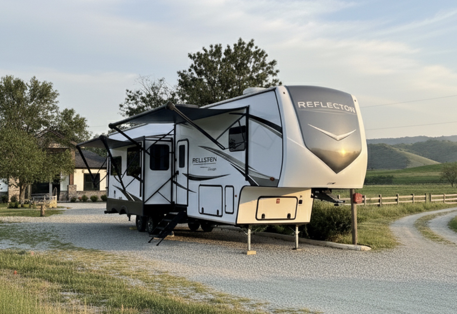 Deluxe fifth-wheel RV with awning extended, parked on a gravel pad in front of a country home at sunset.
